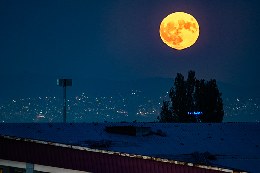 image of the full moon rising over the city center. The moon was photographed with city lights. Shot with a full frame camera and tele lens.