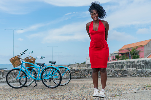 Afro Latina woman with curly hair and black skin is casually dressed in red dress and tennis shoes while riding a bicycle through the historic center of Cartagena enjoying the sunny day