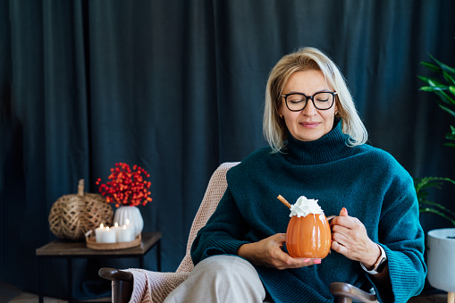 Smiling middle aged woman relaxing with pumpkin shaped cup of hot drink with whipped cream and cinnamon stick with fall mood composition for hygge home decor on background. Cozy autumn holidays.