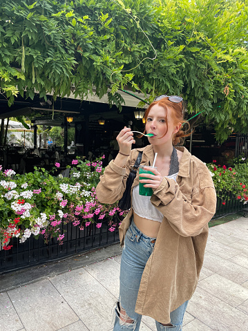 Stock photo showing close-up view of beautiful, red haired, female tourist wearing white crop top, fleece jacket and ripped jeans whilst walking through Venice eating a green, slushie ice drink with a plastic spoon.