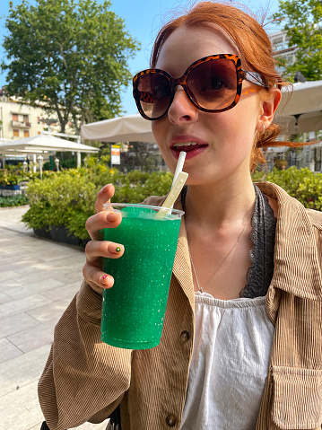 Stock photo showing close-up view of beautiful, red haired, female tourist wearing white crop top, fleece jacket and ripped jeans whilst walking through Venice drinking a green, slushie ice drink with a paper straw.
