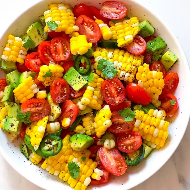 A close-up photo of a corn salad with baby tomatoes, avocado, jalapeño pepper and black pepper in a white bowl.