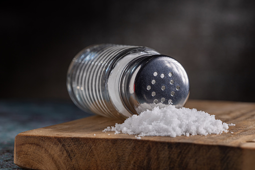 Glass salt shaker shot on rustic wooden table. A spoon with salt is beside the shaker placed directly on the table. The composition is at the left of an horizontal frame leaving useful copy space for text and/or logo at the right. Predominant colors are white and brown. High resolution 42Mp studio digital capture taken with Sony A7rII and Sony FE 90mm f2.8 macro G OSS lens