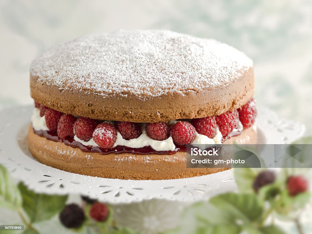 Victoria sponge cake Victoria sponge cake filled with fresh whipped cream, rasberries and jam on white platter. Leafy foiage in the background and foreground out of focus. Shallow depth of field Sponge Cake Stock Photo
