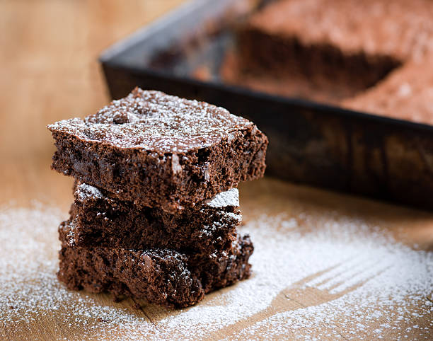 Close-up of stacked pieces of brownies with powdered sugar Newly baked chocolate brownies, stacked three high, with a light dusting of icing sugar. Outline of fork can be seen in the icing on the table. Cake tin with missing squares can be seen out of focus in the background. powdered sugar stock pictures, royalty-free photos & images