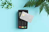 Grey credit card on a portable WiFi contactless card reader on light blue background with plants as decoration. Illustration of the concept of credit card consumerism and its protection