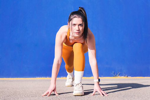 portrait of a focused sportswoman in position to start a race with orange sportswear in a blue background, concept of sport and active lifestyle, copy space for text