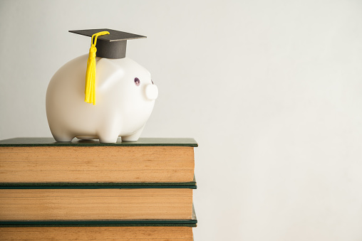 Piggy bank and graduation cap on stack textbooks in classroom with white wall background copy space. Personal financial planning and money management for education, abroad educational, money savings concept.