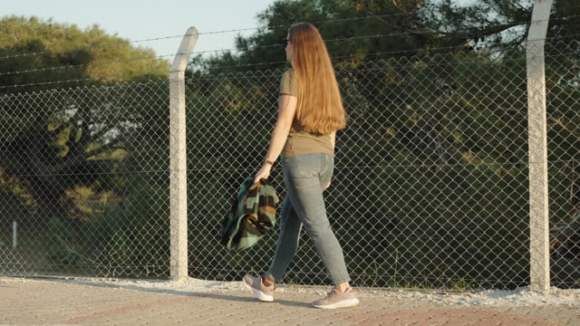 A young woman walks near a forest fenced with a fence made of mesh and barbed wire, sunny clear weather. Slow motion