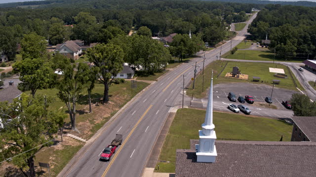 Pickup towing a trailer on a highway passing through Hamilton - a small city in Alabama, on a sunny summer day. Drone aerial footage with forwarding-panning and descending camera motion started from intersection and passing the church.