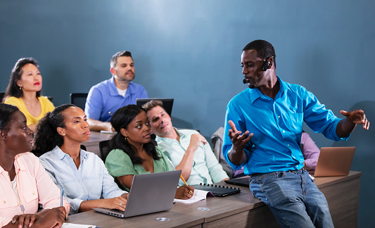 An African-American man in his 30s teaching a multiracial group of adult students who are sitting in rows at long tables. He is in the front of the classroom, using a headset microphone, leaning on one of the tables, looking over his shoulder toward the students as he speaks.
