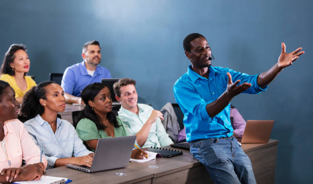 An African-American man in his 30s teaching a multiracial group of adult students who are sitting in rows at long tables. He is in the front of the classroom, using a headset microphone, leaning on one of the table, pointing toward the off-camera visual aid.