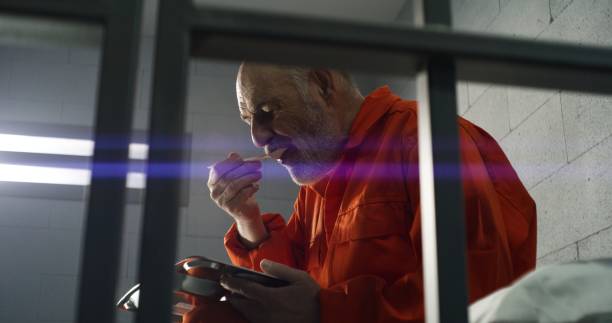 Elderly criminal eats dinner sitting in prison cell in jail Elderly prisoner, criminal in orange uniform eats dinner sitting in prison cell. Offender serves imprisonment term for crime. Jail, detention center or correctional facility. View through metal bars. prison meal stock pictures, royalty-free photos & images
