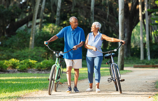 A senior African American couple in the park, walking side by side with their bicycles, conversing. They are looking toward each other. The man has a serious expression, and the woman is smiling.