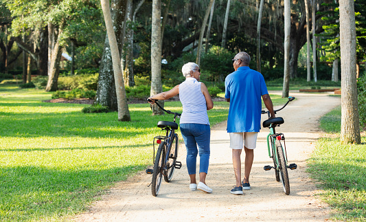 Rear view of a senior African American couple in the park, walking side by side with their bicycles, conversing. They are looking toward each other, smiling.