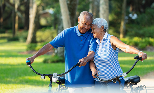 A senior African American couple in the park standing side by side with their bicycles, smiling and affectionately leaning on each other.