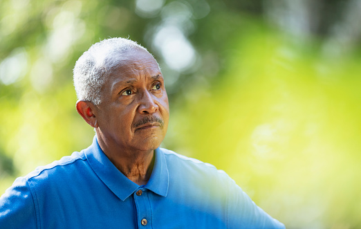 Headshot of a senior African-American man, in his 70s, thinking, hands on his hips. He is outdoors at the park, looking away from the camera, glancing upward, with a serious expression, maybe just a little worried about something.
