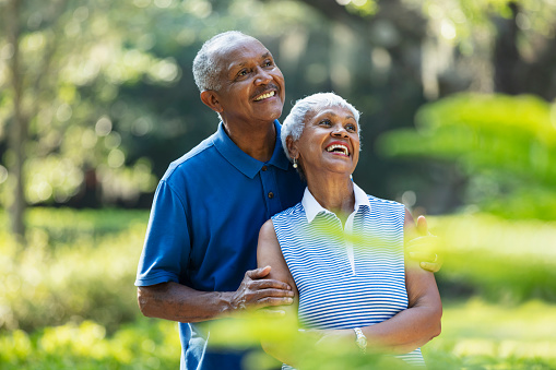 A senior African-American couple, in their 70s, standing together at the park, surrounded by lush foliage, smiling and enjoying the view.
