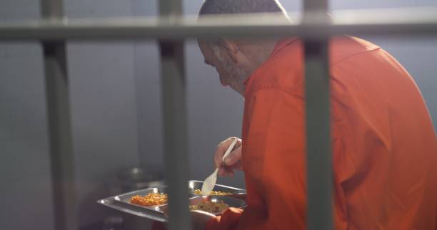 Elderly criminal in orange uniform sits in prison cell, eats dinner Elderly prisoner in orange uniform sits in prison cell, eats dinner. Criminal, inmate serves imprisonment term for crime. Jail, detention center or correctional facility. Shooting through metal bars. prison meal stock pictures, royalty-free photos & images