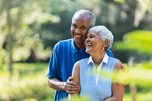 A senior African-American couple, in their 70s, standing together at the park, surrounded by lush foliage. The man is smiling and looking at the camera.