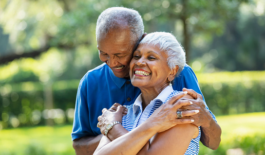 A senior African-American couple, in their 70s, standing together at the park, surrounded by lush foliage. The man is hugging his wife from behind and she is grinning.