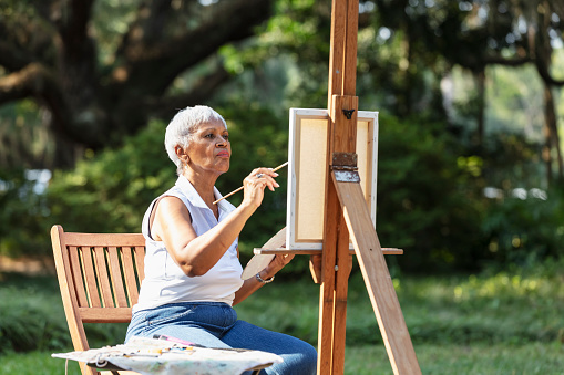 A senior African-American woman painting outdoors, at the park. The artist is working on a small canvas on an easel, with a serious expression.