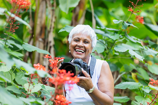 A senior African-American woman, in her 70s, pursuing her photography hobby in retirement. She is at the park with her camera, photographing nature, surrounded by lush foliage. She is laughing and looking at the camera.