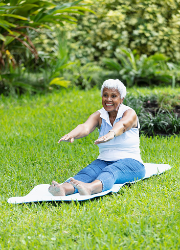 A senior African-American woman, in her 70s, exercising outdoors, sitting on a mat on the grass, stretching. She is smiling, with her arms straight, reaching out in front of her.