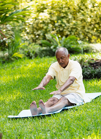 A senior African-American man, in his 70s, exercising outdoors, sitting on a mat on the grass, stretching. He has a serious expression on his face, arms straight out in front, reaching to touch his toes.