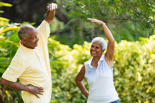 A senior African-American couple exercising together outdoors, stretching. The main focus is on the woman, who is looking at her husband, smiling.