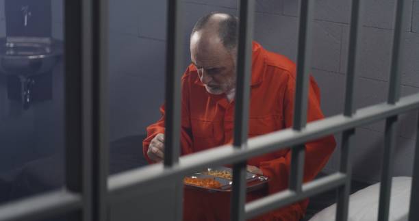 istock Elderly prisoner in orange uniform sits in prison cell, eats food 1617870303