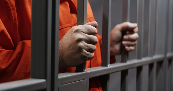 Hands close up of elderly prisoner in orange uniform holding metal bars, standing in prison cell. Criminal serves imprisonment term for crime. Inmate in jail or correctional facility. Justice system.