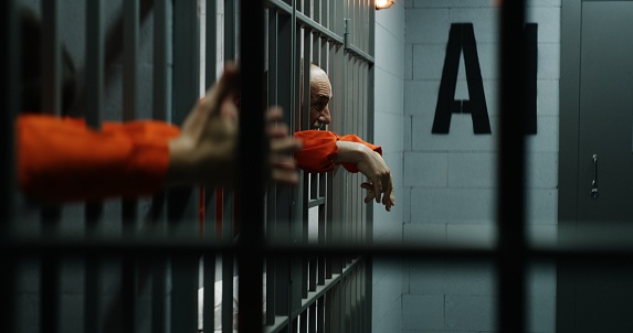Prisoners in orange uniforms lean on metal bars in prison cells. Guilty criminals serve imprisonment terms for crimes in jail, detention center. Elderly inmate speaks aggressively. View from jail cell