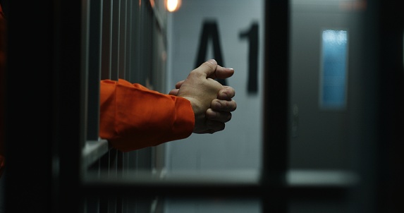 Close up of prisoner in orange uniform holding metal bars, standing in prison cell. Guilty criminal or killer serves imprisonment term for crime. Inmate in jail or detention center. Justice system.