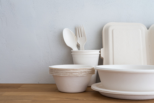 Natural eco-friendly disposable utensils (fork, spoon, dish plate, bowl, cup and fast food box container) made of fiber of bagasse and bamboo on wooden table with white wall background. Save the earth and waste reduction concept.