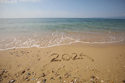 New Year 2024, lettering on the beach with wave and clear blue sea. Numbers 2024 year on the sea shore, message handwritten in the golden sand on beautiful beach background. New Years concept.
