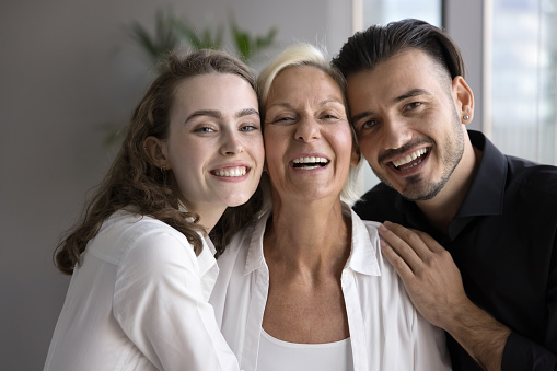 Adult siblings hugging senior mother, standing close with cheek touches, looking at camera with toothy smiles, laughing. Older mom enjoying motherhood, meeting with son and daughter. Family portrait