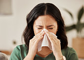 Blowing nose, tissue and woman in a living room with flu, cold and hay fever, crisis or viral infection in her home. Sneezing, allergies and person with virus or coughing illness in a lounge in house