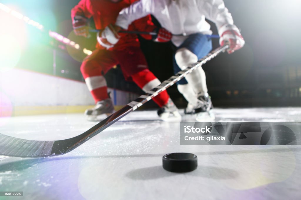 Ice hockey. Ice-level image of male ice hockey players in a tackle during a game. The high white socks and dark blue trousers of the hockey player can be seen.There's a hockey stick reaching for the puck. A hockey puck casting two shadows is on the ice in the right corner of the image. Ice Hockey Stock Photo