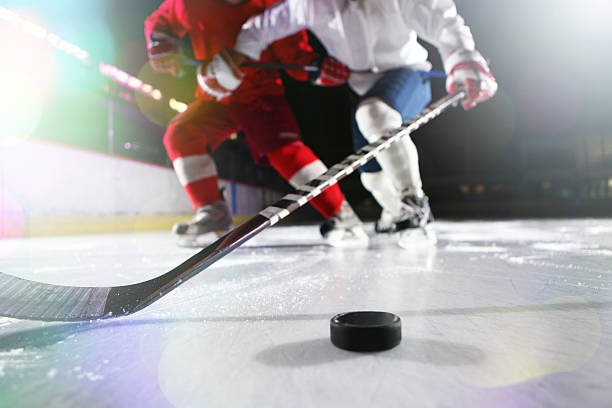 Ice hockey. Ice-level image of male ice hockey players in a tackle during a game. The high white socks and dark blue trousers of the hockey player can be seen.There's a hockey stick reaching for the puck. A hockey puck casting two shadows is on the ice in the right corner of the image. ice hockey stock pictures, royalty-free photos & images