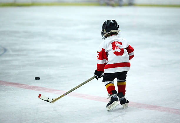 Junior ice hockey. Rear view of unrecognizable 6 year old boy at ice hockey practice trying to reach the puck. He's wearing black helmet and white and red jersey and red socks.  hockey stock pictures, royalty-free photos & images