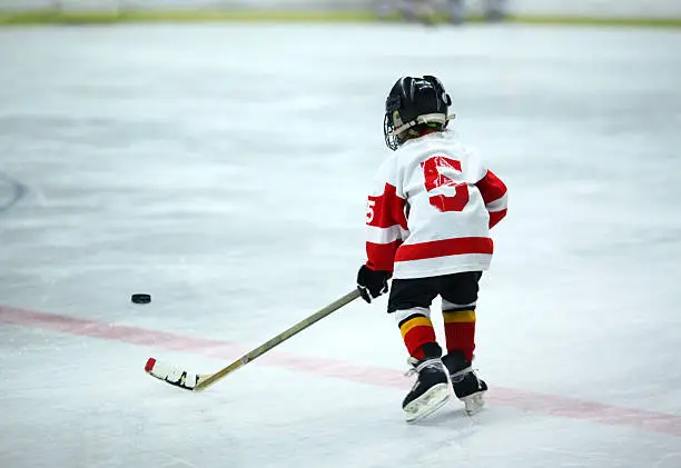 Rear view of unrecognizable 6 year old boy at ice hockey practice trying to reach the puck. He's wearing black helmet and white and red jersey and red socks. 