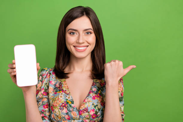 istock Photo of charming positive lady demonstrate empty space smart phone display direct finger offer isolated on green color background 1618206749