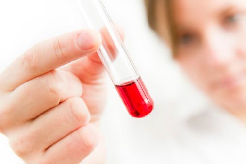 Female researcher analysing test tube filled with red substance. Focus on foreground: Hand with test tube. Face blurry in background.