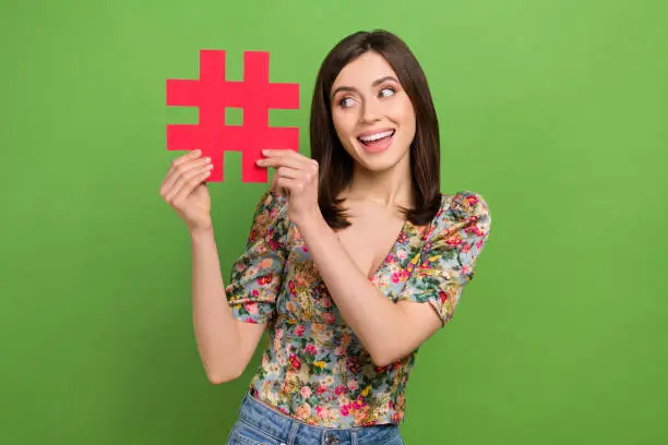 Photo of Photo of young funky blogging woman hold paper icon hash tag social media popularity advert instagram isolated on green color background