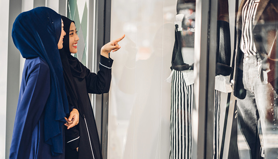 Two young asian muslim woman enjoying shopping and having fun pointing finger in the shop window outdoors at fashion store