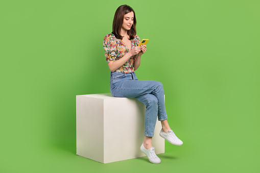 Full size photo of pretty girl sit cube podium use smart phone chatting isolated on green color background.