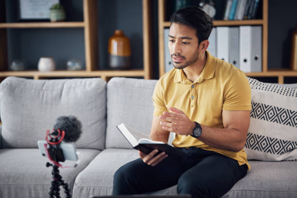istock Podcast, bible study and phone with a man online to preach or reading while live streaming. Asian male on home sofa with Christian scripture or book as content creator teaching on education religion 1618249537