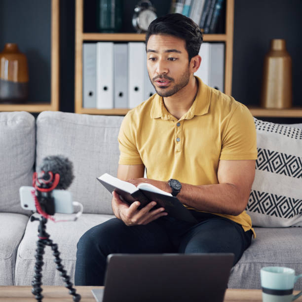 istock Podcast, bible and man reading with phone and microphone online while live streaming. Asian male on home sofa with Christian religion book as blog content creator or influencer teaching or studying 1618249538