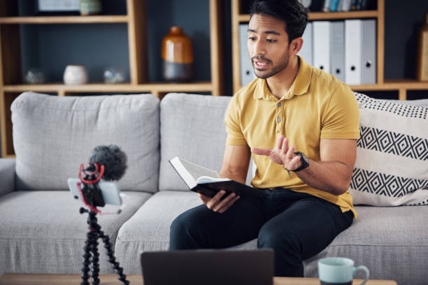 istock Book, bible study and phone with a man online to preach or reading while live streaming. Asian male on home sofa talking Christian scripture as podcast content creator teaching education on religion 1618250002